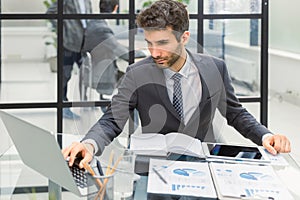 Young modern business man analyzing data using laptop while working in the office with collegues on the background.