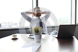 Young modern business man keeping hands behind head and smiling while sitting in the office