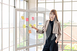 young modern businesswoman in casual fashion suit looking at the camera while using sticky notes for strategy ideas on glass board