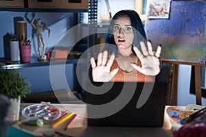 Young modern girl with blue hair sitting at art studio with laptop at night afraid and terrified with fear expression stop gesture