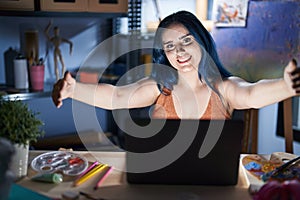 Young modern girl with blue hair sitting at art studio with laptop at night looking at the camera smiling with open arms for hug