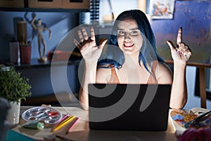 Young modern girl with blue hair sitting at art studio with laptop at night showing and pointing up with fingers number seven