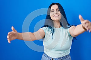 Young modern girl with blue hair standing over blue background looking at the camera smiling with open arms for hug