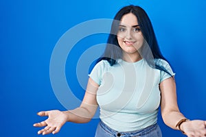 Young modern girl with blue hair standing over blue background smiling cheerful with open arms as friendly welcome, positive and