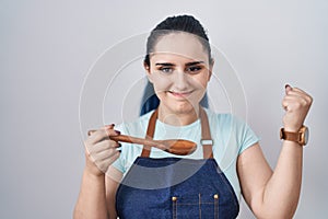 Young modern girl with blue hair wearing cook apron holding spoon screaming proud, celebrating victory and success very excited