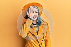 Young modern girl wearing yellow hat and leather jacket covering one eye with hand, confident smile on face and surprise emotion