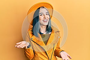Young modern girl wearing yellow hat and leather jacket smiling cheerful with open arms as friendly welcome, positive and