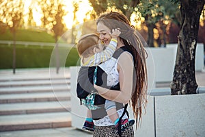 Young happy mother with baby son in ergo backpack walking in Sunny summer day. Concept of the joy of motherhood