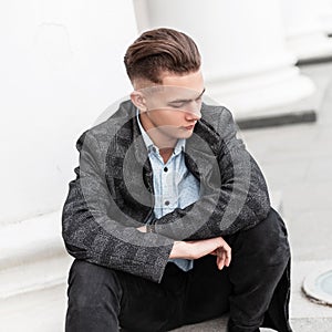 Young modern man in elegant checkered gray jacket in a stylish shirt in trendy jeans with a fashionable hairstyle is resting