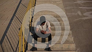 Young modern man without shoes, sitting on urban stairs, using smartphone, wearing black cap and socks.