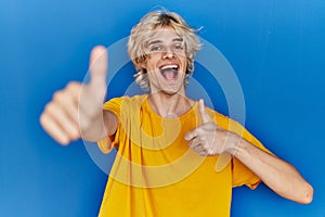 Young modern man standing over blue background approving doing positive gesture with hand, thumbs up smiling and happy for success