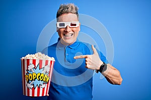 Young modern man watching cinema movie wearing 3d glasses and eating popcorn with surprise face pointing finger to himself