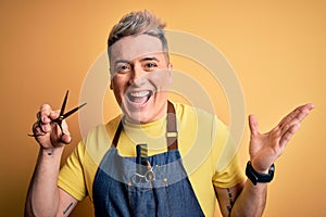 Young modern professional hairdresser man holding scissors over isolated yellow background very happy and excited, winner