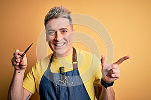 Young modern professional hairdresser man holding scissors over isolated yellow background very happy pointing with hand and