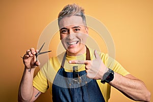 Young modern professional hairdresser man holding scissors over isolated yellow background very happy pointing with hand and