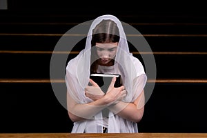 A young modest girl with a handkerchief on her head and a bible in her hands is sitting in church and praying. The concept of