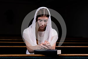 A young modest girl with a handkerchief on her head and a bible in her hands is sitting in church and praying. The concept of