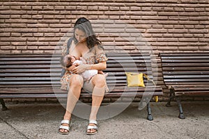 Young mom breastfeeding newborn baby daughter in public outdoors in the street on a bench