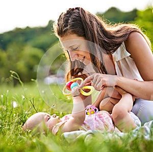 Young mom and her baby daughter playing together with a rattle in the grass outddors - Carefree childhood and family concept