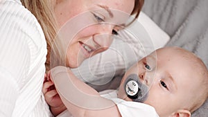 Young mom hugs and kisses infant with pacifier while lying on cozy bed, close-up. Mother and her newborn baby enjoy