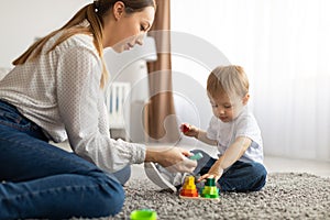Young mom and toddler child playing together at home, using toys while sitting on floor carpet in living room