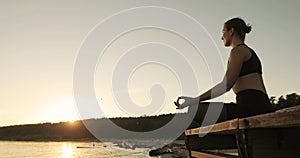 Young moman is meditating sitting on the river pier at sunset in lotus pose.