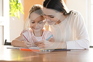 Young mommy embracing shoulders of happy small daughter, drawing pictures.