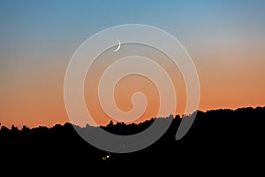 A young moon on an orange-blue sunset sky over a dark forest. Horizontal orientation. High quality photo
