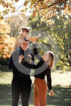 Young mother and adorable little daughter sitting on fathers shoulders are walking outdoors at autumn park. Dad, mom and