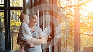 Young mother approaches window holding baby and smiling