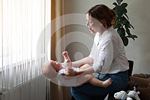 Young mother and baby daughter, son are having fun at home by window together. Beautiful mom throws her adorable newborn