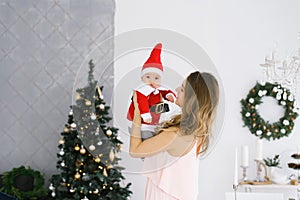 A young mother with a baby in a little Santa costume in her arms near the Christmas tree in the living room