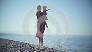 Young mother with a baby in their hands walking on the beach. The baby is smiling and waving at the camera.