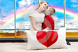 A young mother with a beautiful baby son holding a pillow in shape of heart on the background of the panoramic Windows with a rain