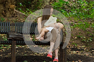 Young mother breastfeeding baby in the park. Mom sits on bench and holds tiny tot in arms
