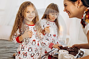 Young mother is bringing cocoa with Marshmallows and cookies to her daughters in pajamas sitting on the bed in the full