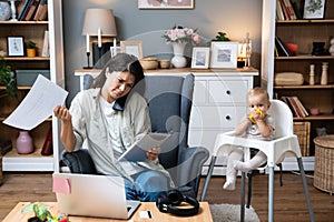 Young mother, business woman having cellphone conversation from home while taking care of her baby sitting in tall baby chair.