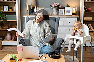 Young mother, business woman having cellphone conversation from home while taking care of her baby sitting in tall baby chair.