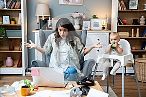 Young mother, business woman having video call on laptop from home while taking care of her baby sitting in tall baby chair.