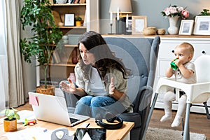 Young mother, business woman having video call on laptop from home while taking care of her baby sitting in tall baby chair.