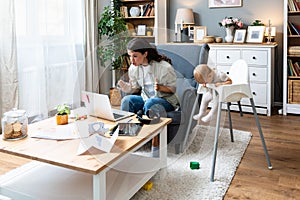 Young mother, business woman having video call on laptop from home while taking care of her baby sitting in tall baby chair.