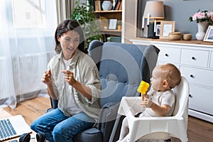 Young mother, business woman preparing for work from home while taking care of her baby sitting in tall baby chair. Female