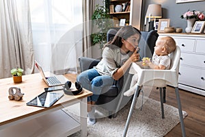 Young mother, business woman preparing for work from home while taking care of her baby sitting in tall baby chair. Female