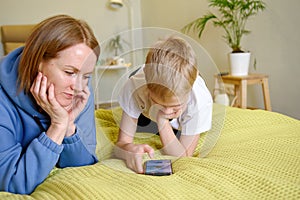 Young mother in casual clothes is lying on the bed next to her son, looking at information on the phone. Happy children with their