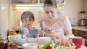 Young mother checking recipe on smartphone while baking pie or cake with her little son. Children cooking with parents