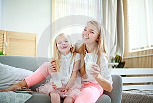 Young mother with child daughter at home on couch drinking milk and cookies, laughing and talking together