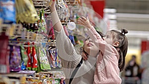 Young mother and child near the shelves with sweets in the supermarket. Little girl chooses candy in brightly colored