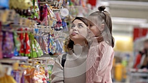 Young mother and child near the shelves with sweets in the supermarket. Little girl chooses candy in brightly colored