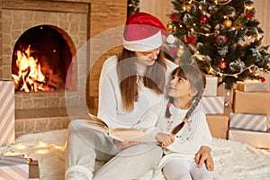 Young mother and child reading book on winter evening by fireplace, mom and female kid looking at each other with love, wearing