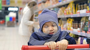 Young mother is choosing food in supermarket on the background of very sad 3 years old boy sitting in the trolley